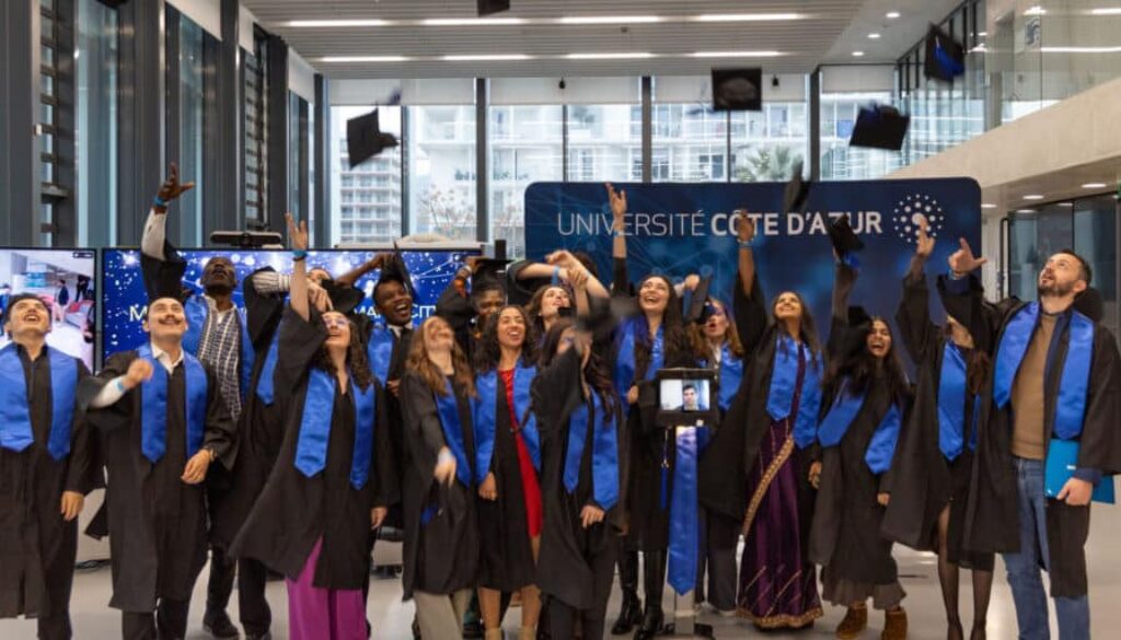 Des diplômés en toge noire et écharpe bleue célèbrent leur remise de diplômes à l’Université Côte d’Azur, lançant joyeusement leurs bonnets en l’air. Un étudiant participe à distance via un robot de téléprésence, visible au centre du groupe.