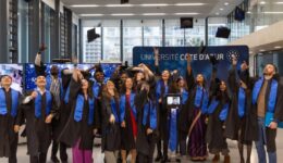 Des diplômés en toge noire et écharpe bleue célèbrent leur remise de diplômes à l’Université Côte d’Azur, lançant joyeusement leurs bonnets en l’air. Un étudiant participe à distance via un robot de téléprésence, visible au centre du groupe.