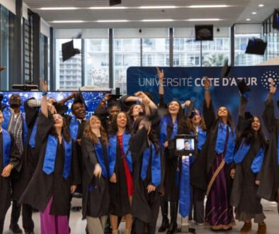 Des diplômés en toge noire et écharpe bleue célèbrent leur remise de diplômes à l’Université Côte d’Azur, lançant joyeusement leurs bonnets en l’air. Un étudiant participe à distance via un robot de téléprésence, visible au centre du groupe.